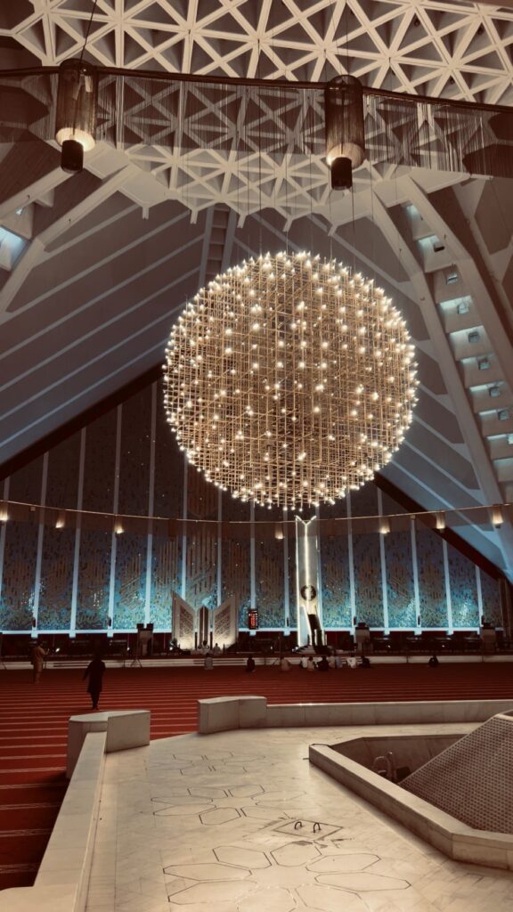 Interior view of Faisal Mosque’s main prayer hall featuring a massive, glowing spherical chandelier under a geometric ceiling with warm ambient lighting.