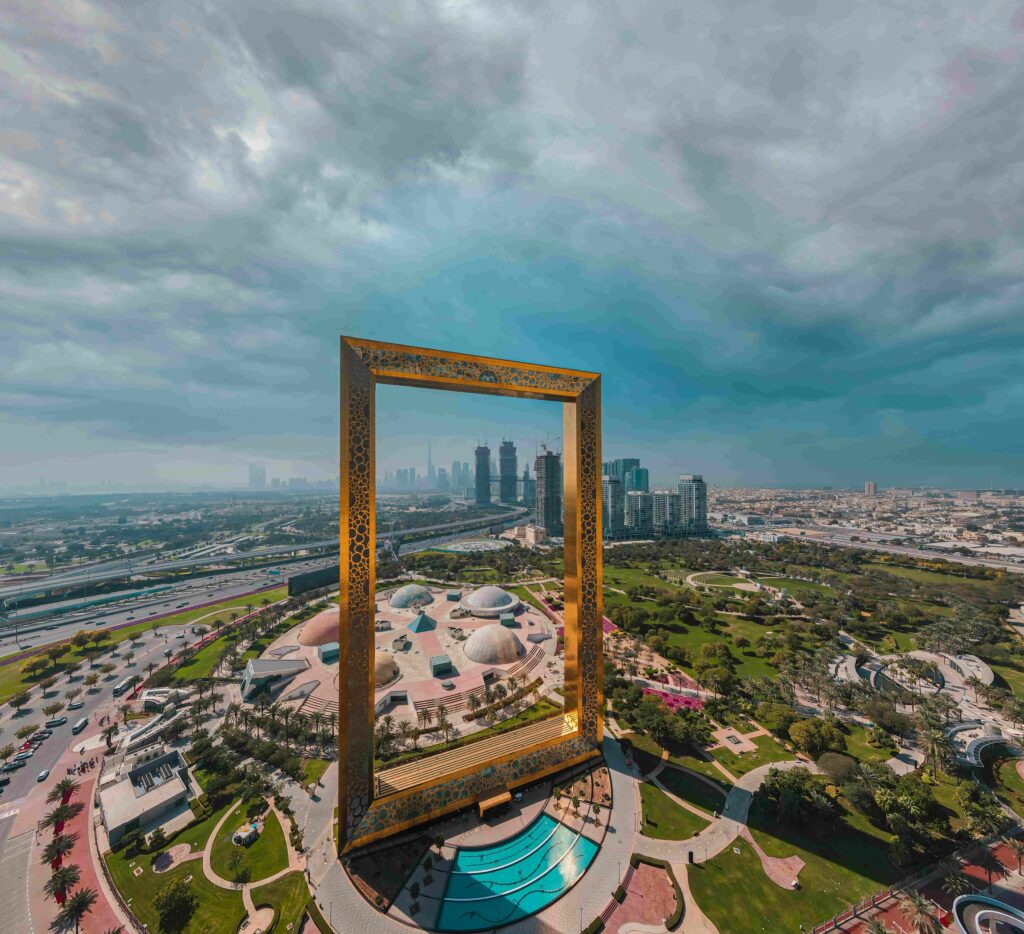 Aerial drone view of the Dubai Frame standing in Zabeel Park with the modern Dubai skyline including the Burj Khalifa visible through its golden opening, under dramatic storm clouds