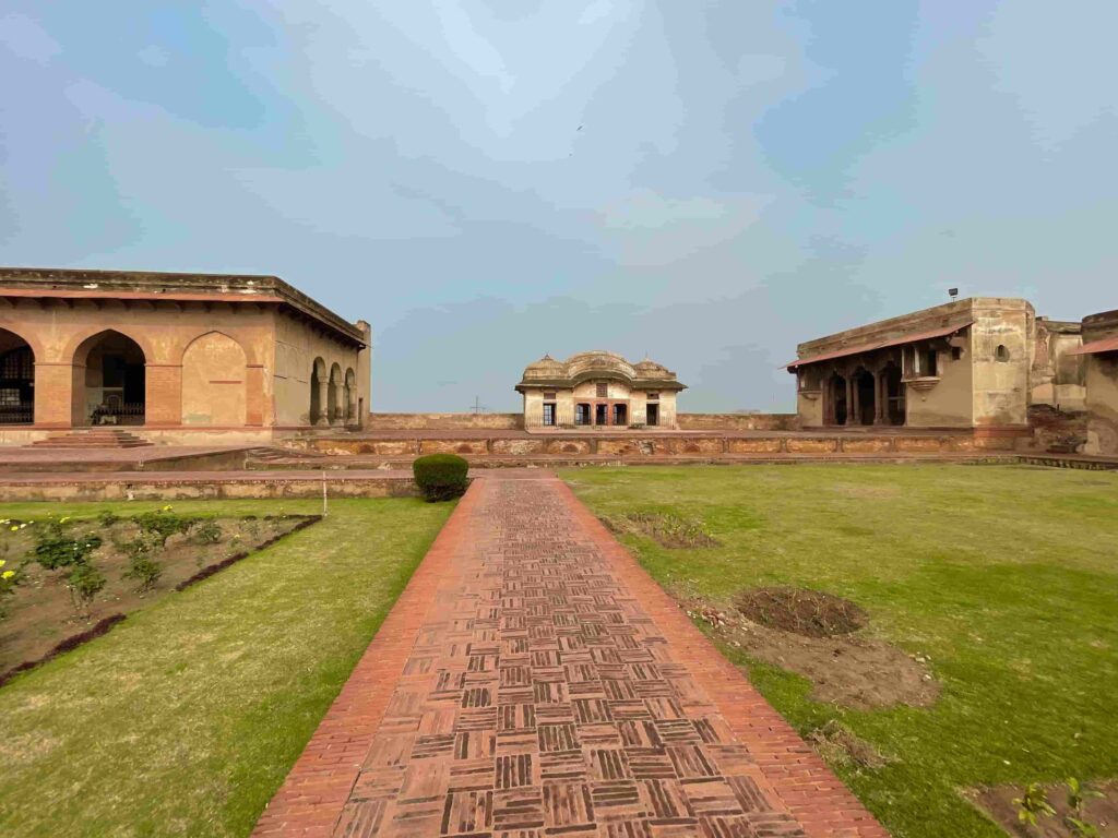 A wide-angle view of the brick walkway and green lawns leading to the historic Diwan-i-Khas buildings inside Lahore Fort under an overcast sky.