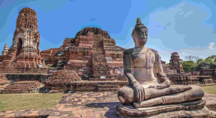 Ancient stone Buddha statue sitting in front of historic temple ruins in Ayutthaya Thailand under a clear blue sky