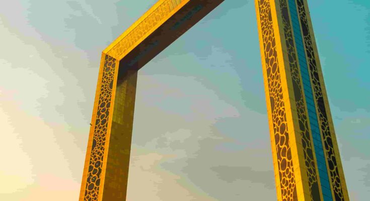 The Dubai Frame photographed from below at golden hour, its gold-latticed towers rising against a teal and amber sky with palm trees in the foreground, Dubai, UAE