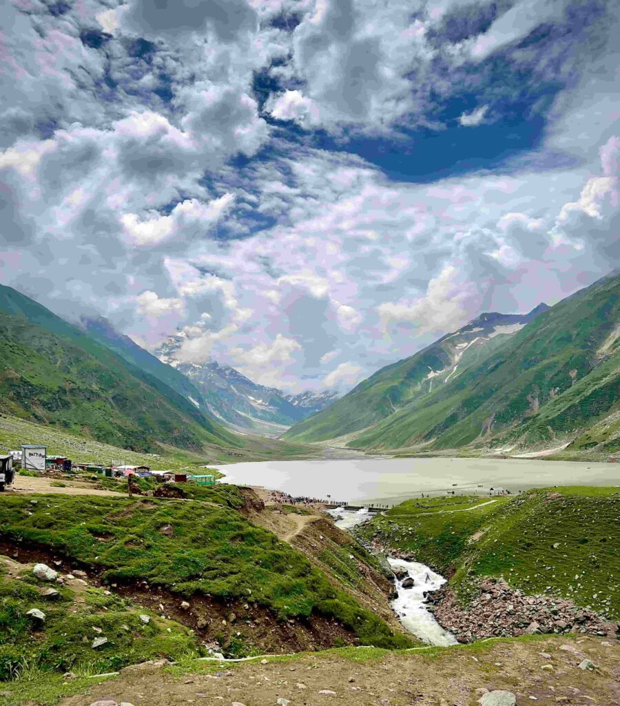 A high-angle view of the iconic Lake Saif-ul-Malook nestled between steep green mountains under a dramatic, cloudy sky. A narrow stream of water flows out from the lake into the valley, and small tourist stalls are visible on the grassy slopes in the foreground.