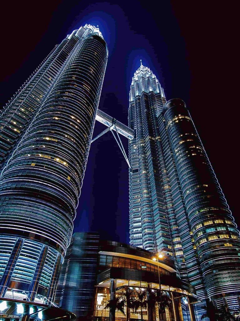 Low angle view of Petronas Twin Towers glowing blue at night with Sky Bridge visible in Kuala Lumpur