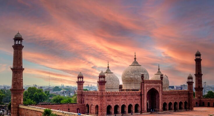 Majestic panoramic view of Badshahi Mosque Lahore at sunset, showcasing its red sandstone structure and white marble domes under a vibrant orange sky.