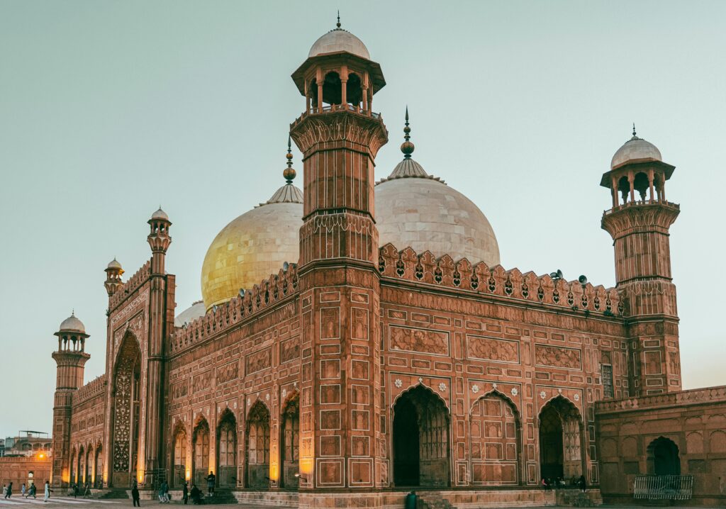 A stunning low-angle view of the Badshahi Mosque in Lahore at dusk, showcasing its intricate red sandstone architecture, grand domes, and minarets against a soft evening sky.