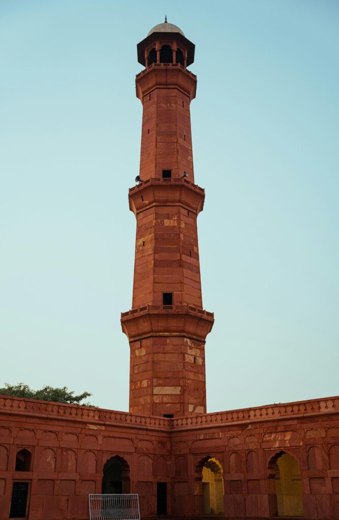 A towering red sandstone minaret of the Badshahi Mosque in Lahore, shot from a low angle against a clear, pale blue sky, highlighting the intricate Mughal architectural details.