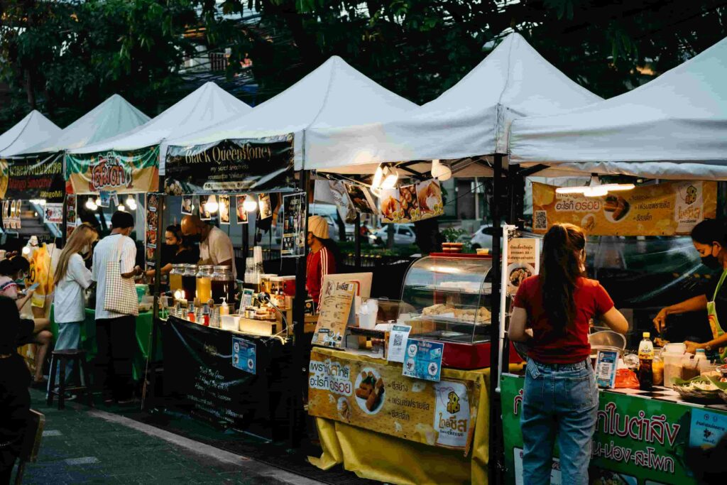 Local street food vendors preparing snacks under illuminated white tents at an outdoor night market in Bangkok