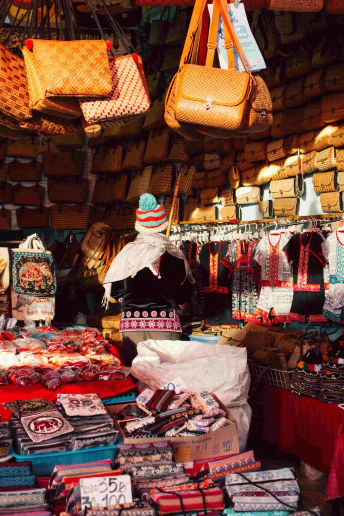 A colorful Thai market stall in Chiang Mai, specializing in woven bamboo bags and traditional textile clothing, with a vendor seen from behind wearing a colorful hat.
