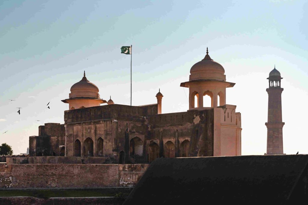 A cinematic side view of the massive red brick walls of Lahore Fort and a distant minaret, showcasing the grand Mughal architecture during a hazy sunset.