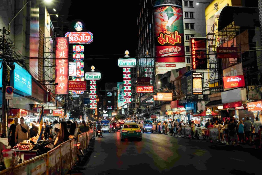 Bright neon signs in Chinese and Thai hanging over a crowded Yaowarat Road street market in Bangkok Chinatown at night