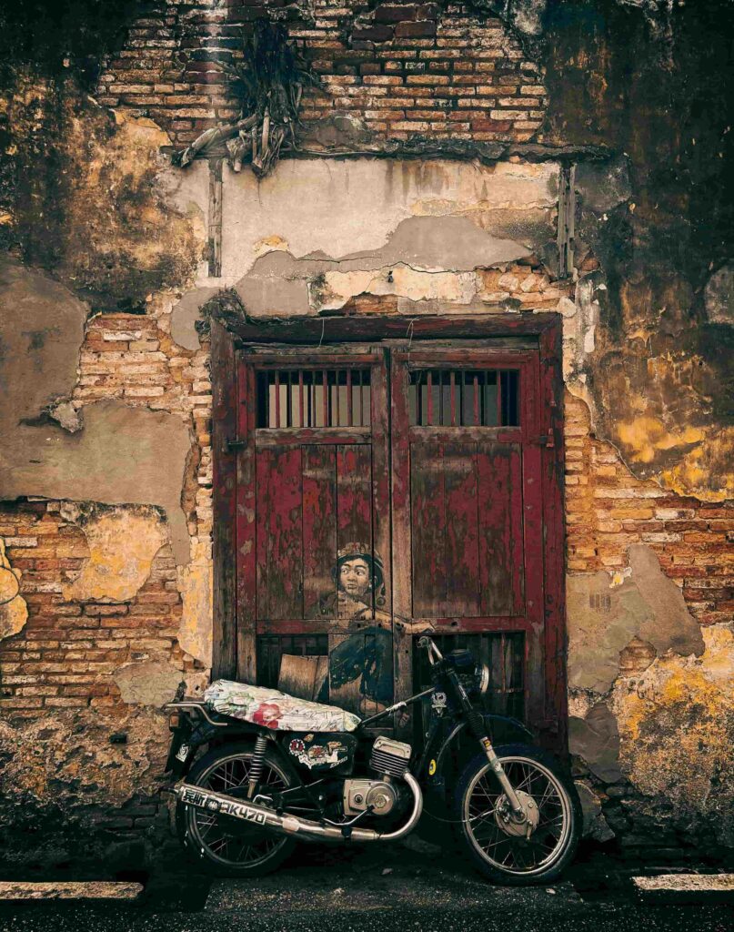 Vintage motorcycle parked in front of a weathered wall with a faded street art mural on a rustic red door in George Town, Penang
