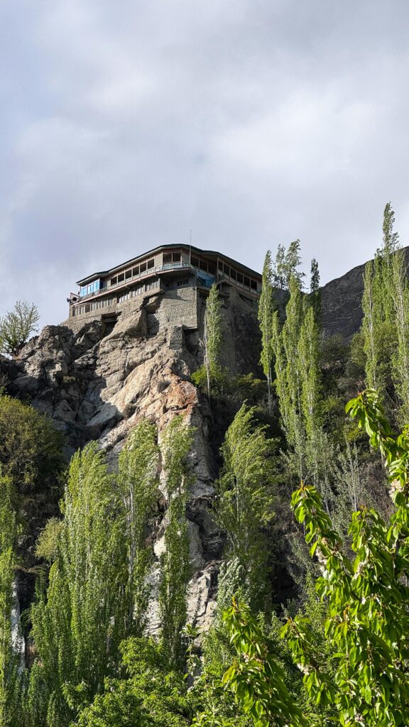 "A low-angle shot of the historic Baltit Fort in Hunza Valley, Pakistan, perched on a rocky cliff and surrounded by tall green trees under a cloudy sky."