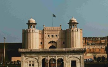 Alt Text: A symmetrical front view of the white marble Naulakha Pavilion at Lahore Fort, with the Pakistani flag flying over the Alamgiri Gate towers in the background under a clear sky.