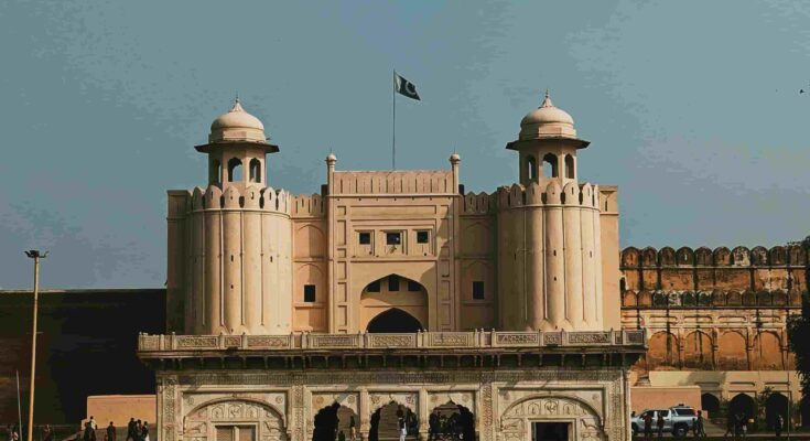 Alt Text: A symmetrical front view of the white marble Naulakha Pavilion at Lahore Fort, with the Pakistani flag flying over the Alamgiri Gate towers in the background under a clear sky.