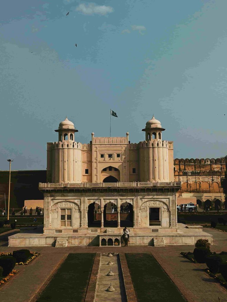 Alt Text: A symmetrical front view of the white marble Naulakha Pavilion at Lahore Fort, with the Pakistani flag flying over the Alamgiri Gate towers in the background under a clear sky. Pakistan 7 Days Budget Travel Plan