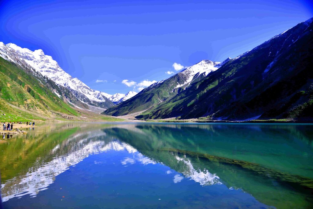 A wide landscape view of Lake Lulusar with crystal-clear turquoise water. The calm surface of the lake perfectly reflects the surrounding snow-capped mountains and the deep blue sky. A few small figures of tourists can be seen on the rocky shore to the left.