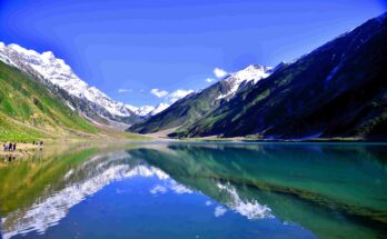 A wide landscape view of Lake Lulusar with crystal-clear turquoise water. The calm surface of the lake perfectly reflects the surrounding snow-capped mountains and the deep blue sky. A few small figures of tourists can be seen on the rocky shore to the left.