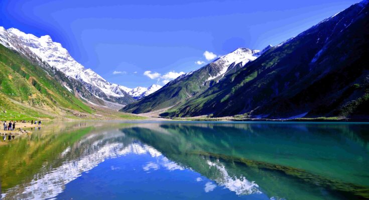 A wide landscape view of Lake Lulusar with crystal-clear turquoise water. The calm surface of the lake perfectly reflects the surrounding snow-capped mountains and the deep blue sky. A few small figures of tourists can be seen on the rocky shore to the left.