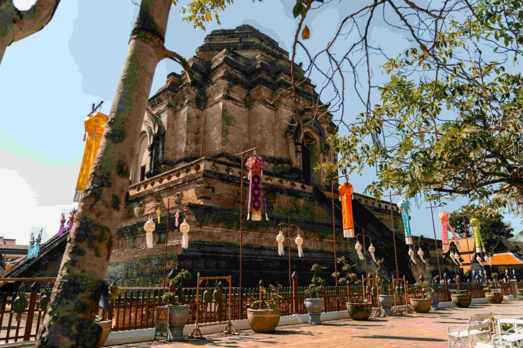 The majestic, weathered brick ruins of Wat Chedi Luang in Chiang Mai, viewed under a clear blue sky through the natural frame of tree branches.