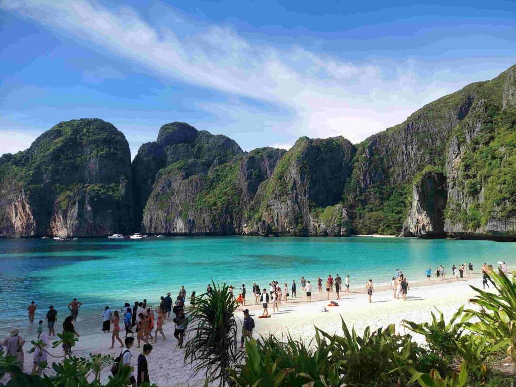 International tourists walking on the white sands of Maya Bay under a bright sky with tropical cliffs
