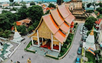 An epic aerial drone photograph looking down upon the complete monastery complex of Wat Chedi Luang in Chiang Mai, with its bright orange roofed temple and ancient ruins surrounded by the modern city.