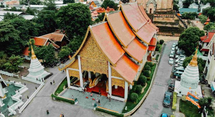 An epic aerial drone photograph looking down upon the complete monastery complex of Wat Chedi Luang in Chiang Mai, with its bright orange roofed temple and ancient ruins surrounded by the modern city.