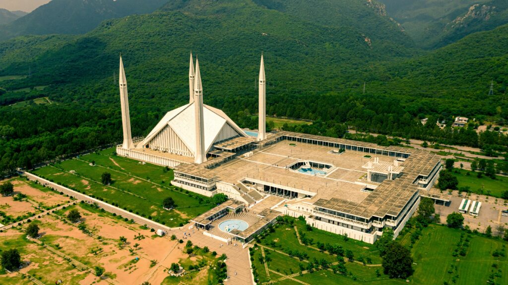 An aerial drone view of Faisal Mosque in Islamabad, showcasing its unique tent-shaped white architecture and four tall minarets against the lush green Margalla Hills.