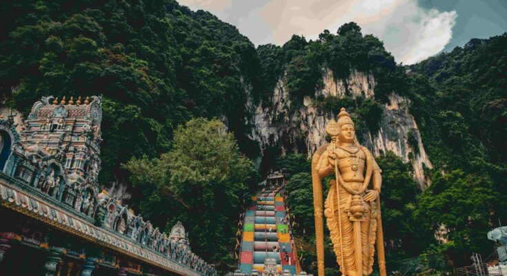 Golden Lord Murugan statue standing in front of rainbow stairs and limestone cliffs at Batu Caves Kuala Lumpur Malaysia