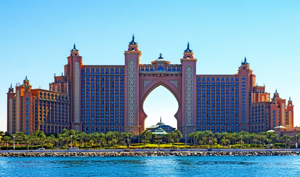 A front-facing wide shot of the Atlantis, The Palm resort in Dubai, featuring its distinct pink Arabian-style architecture and central archway situated along the turquoise waters of the Arabian Gulf under a clear blue sky.