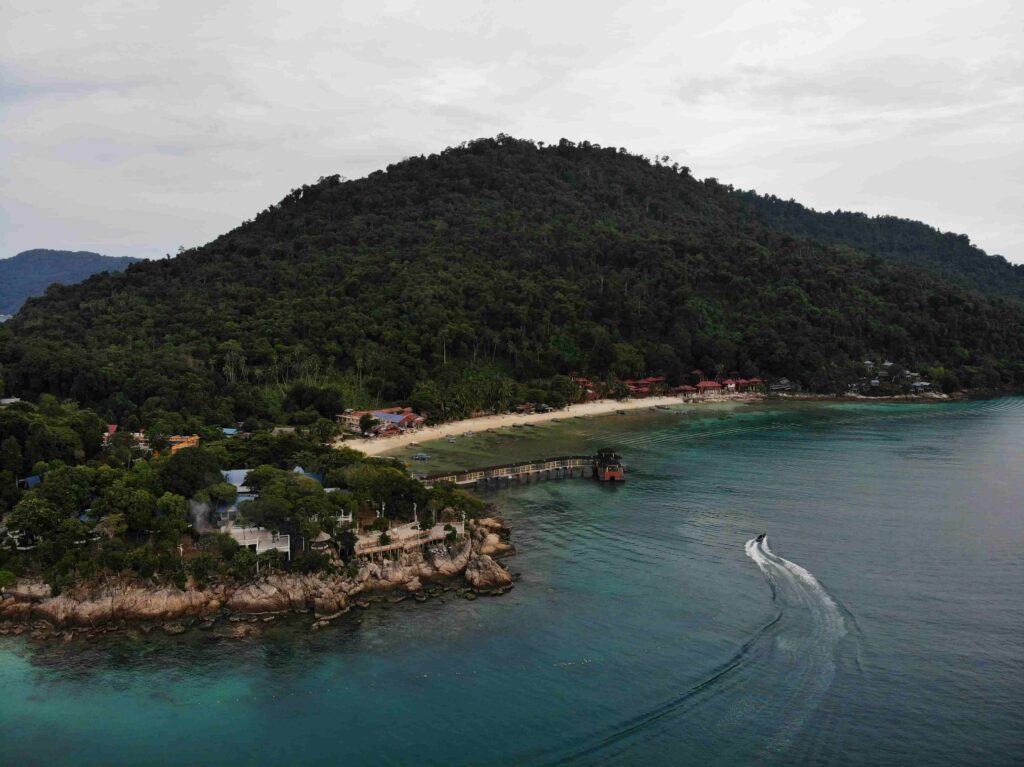 Drone shot of Perhentian Kecil island coastline with a speedboat cutting through teal waters, a sandy beach, jungle-covered hills, and a wooden jetty in the foreground