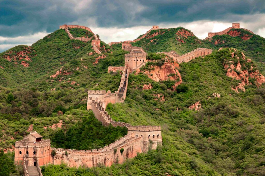 Panoramic view of the Great Wall of China with multiple watchtowers stretching across green mountains under dramatic cloudy sky at Jinshanling section