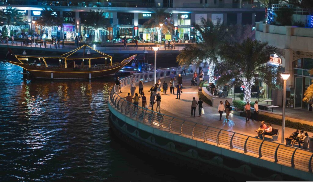 People walking along Dubai Marina promenade at night with a dhow boat docked nearby. Dubai Marina Dhow Cruise Dinner
